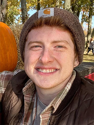 Alex Burks. A smiling individual with short red hair and stubble, wearing a brown beanie hat and earth toned flannel shirt.